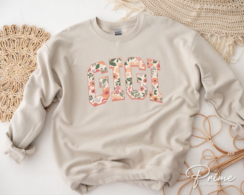 May include: A beige sweatshirt with the word "GIGI" printed in large, bold letters. The letters are filled with a floral pattern in shades of pink, orange, and green.