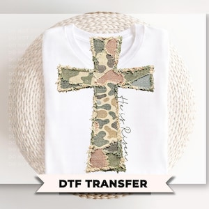 May include: White t-shirt featuring a camouflage cross design. The cross is made of various fabric patches in shades of green, brown, and tan. The words "He is Risen" are written in a script font. The bottom of the image has the text "DTF TRANSFER".