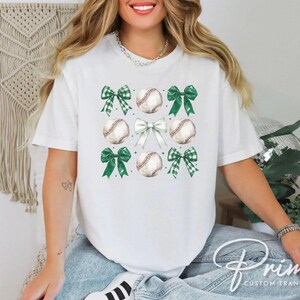 May include: White t-shirt with a design of green and white bows and baseballs. The design is centered on the shirt.