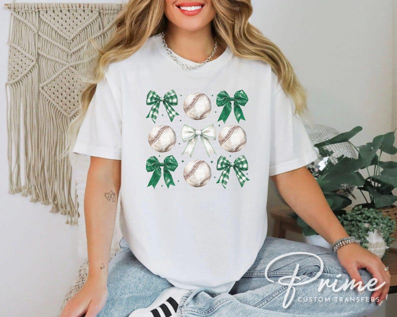 May include: White t-shirt with a design of green and white bows and baseballs. The design is centered on the shirt.
