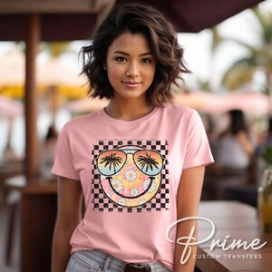May include: A pink t-shirt with a checkered background and a smiley face graphic. The smiley face is wearing sunglasses with palm trees reflected in the lenses. The graphic is surrounded by daisies.