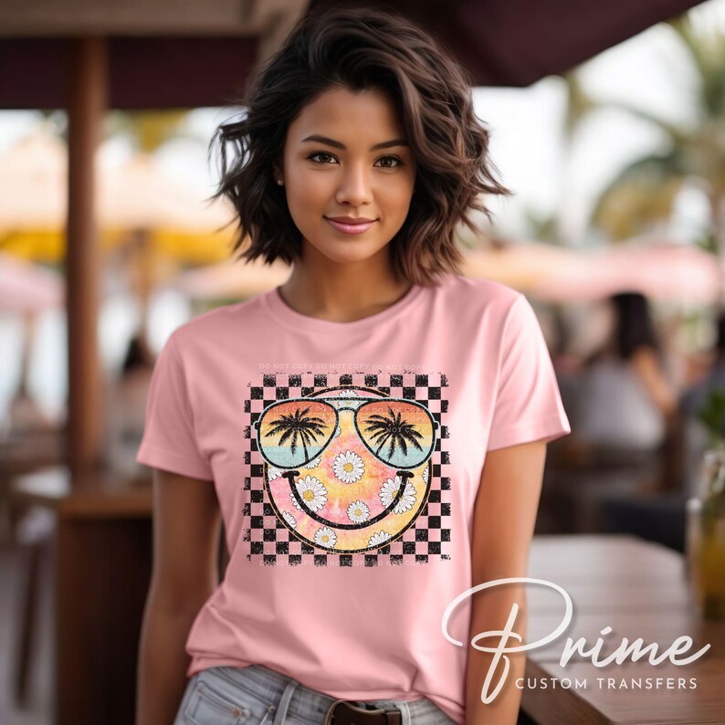 May include: A pink t-shirt with a checkered background and a smiley face graphic. The smiley face is wearing sunglasses with palm trees reflected in the lenses. The graphic is surrounded by daisies.