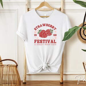 May include: White t-shirt with a red and pink "Strawberry Festival" graphic. The design includes two strawberries, the years "1968", and the phrase "A Berry Good Time". The shirt is hanging on a wooden rack.