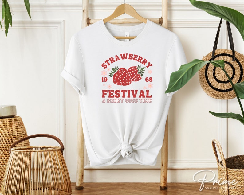 May include: White t-shirt with a red and pink "Strawberry Festival" graphic. The design includes two strawberries, the years "1968", and the phrase "A Berry Good Time". The shirt is hanging on a wooden rack.