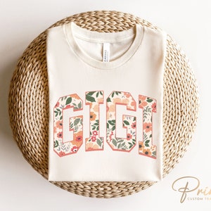May include: A white t-shirt with the word "GIGI" printed in a floral pattern. The letters are outlined in black and the flowers are in shades of pink, orange, and green.