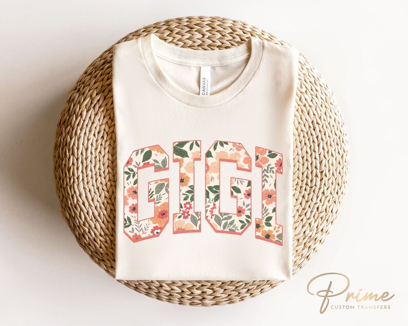 May include: A white t-shirt with the word "GIGI" printed in a floral pattern. The letters are outlined in black and the flowers are in shades of pink, orange, and green.