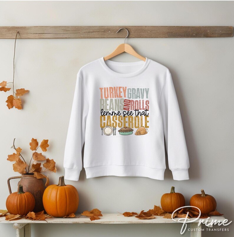 May include: White sweatshirt with a Thanksgiving-themed graphic that says "Turkey Gravy Beans Rolls Lemme See That Casserole". The graphic is in a retro style with colorful text and illustrations of a casserole dish, a turkey, and a roll.