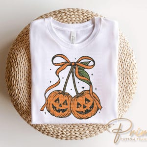 May include: White t-shirt featuring a Halloween-themed design. The graphic depicts two orange pumpkins with carved faces, connected by stems and a bow. The pumpkins have a vintage, distressed look, with small stars around them.