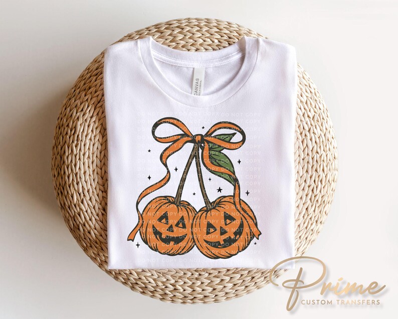 May include: White t-shirt featuring a Halloween-themed design. The graphic depicts two orange pumpkins with carved faces, connected by stems and a bow. The pumpkins have a vintage, distressed look, with small stars around them.