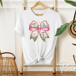 K&ouml;nnte beinhalten: Wei&szlig;es T-Shirt mit einem rosafarbenen Schleifendesign. Die Schleife ist mit Bildern eines Kreuzes, L&auml;mmern und Engeln gef&uuml;llt. Der Text "Prime Custom Transfers" ist in der rechten unteren Ecke sichtbar.