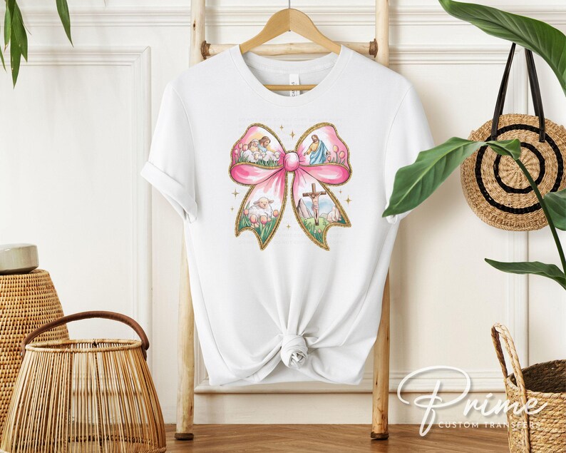 K&ouml;nnte beinhalten: Wei&szlig;es T-Shirt mit einem rosafarbenen Schleifendesign. Die Schleife ist mit Bildern eines Kreuzes, L&auml;mmern und Engeln gef&uuml;llt. Der Text "Prime Custom Transfers" ist in der rechten unteren Ecke sichtbar.