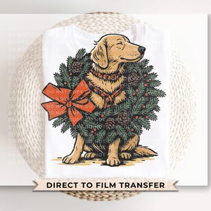 May include: White t-shirt featuring a golden retriever dog wearing a Christmas wreath with a red bow. The dog is sitting, and the wreath includes pinecones and berries. The text "DIRECT TO FILM TRANSFER" is at the bottom.