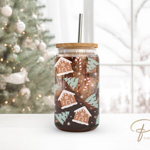 Op de afbeelding: Een transparante glazen tumbler met een bamboe deksel en een roestvrijstalen rietje. De tumbler is versierd met een feestelijk kerstontwerp met gingerbread houses, snoepstokken en groenblijvende bomen. De tumbler is gevuld met een donkerbruine vloeistof.