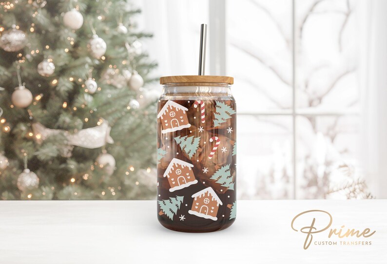 Op de afbeelding: Een transparante glazen tumbler met een bamboe deksel en een roestvrijstalen rietje. De tumbler is versierd met een feestelijk kerstontwerp met gingerbread houses, snoepstokken en groenblijvende bomen. De tumbler is gevuld met een donkerbruine vloeistof.