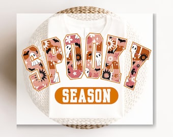 May include: White t-shirt with a graphic of the word "Spooky" in orange and pink letters with a pattern of ghosts, bats, and stars. The text "Season" is below the word "Spooky" in a smaller orange oval.