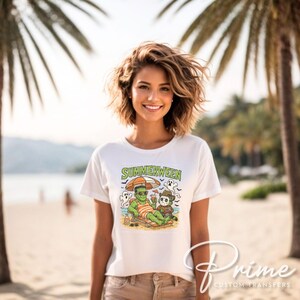 May include: White t-shirt with a colorful graphic featuring Frankenstein and a skeleton enjoying a beach vacation under an umbrella. The text "SUMMERWEEN" is above the graphic. The shirt is worn by a person on a beach.