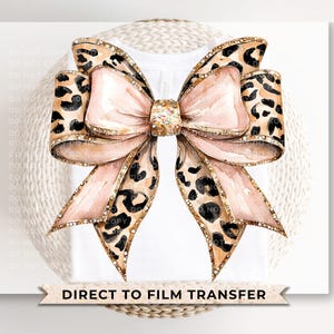 May include: A large pink bow with a leopard print pattern and gold accents. The bow is on a white background with the text "DIRECT TO FILM TRANSFER".