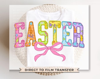 May include: A white t-shirt with a colorful Easter themed design. The design features the word "Easter" in a patchwork style with bunnies and eggs. A pink bow is centered below the word.