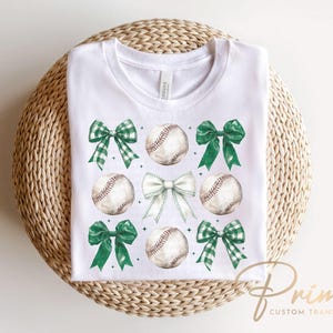 May include: White t-shirt with a green, white, and brown baseball and bow design. The design features four baseballs with green bows and white bows.