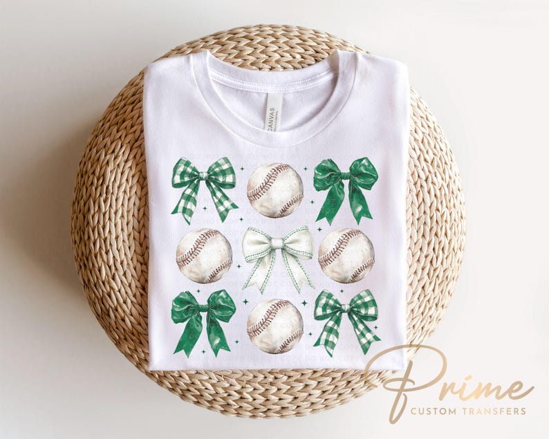 May include: White t-shirt with a green, white, and brown baseball and bow design. The design features four baseballs with green bows and white bows.