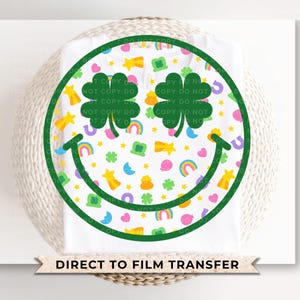 May include: White t-shirt featuring a green smiley face design with shamrocks, rainbows, and stars. The smiley face is outlined in green and filled with colorful St. Patrick's Day themed graphics. The text "DIRECT TO FILM TRANSFER" is at the bottom.