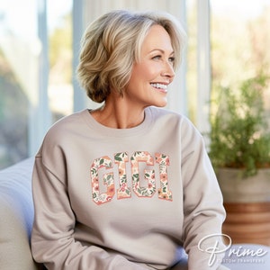 May include: A light beige sweatshirt with a floral print design that spells out the word "GIGI" in a varsity style font.
