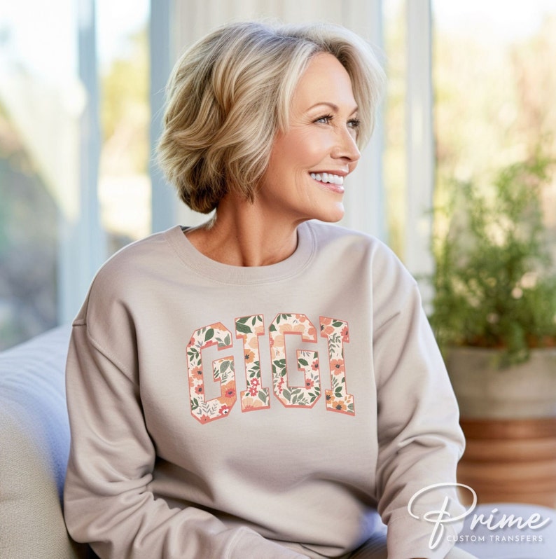 May include: A light beige sweatshirt with a floral print design that spells out the word "GIGI" in a varsity style font.