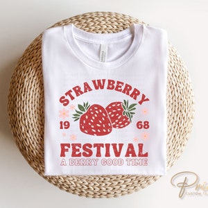 May include: White t-shirt with a retro design featuring the words "Strawberry Festival" above two red strawberries. The text "A Berry Good Time" is below the strawberries. The shirt is folded on a woven surface.