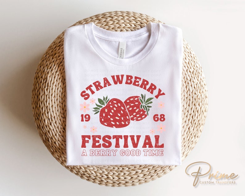 May include: White t-shirt with a retro design featuring the words "Strawberry Festival" above two red strawberries. The text "A Berry Good Time" is below the strawberries. The shirt is folded on a woven surface.
