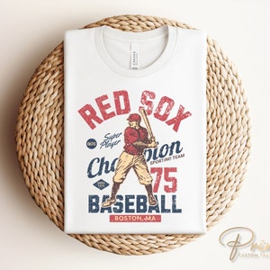 Może przedstawiać: Biała koszulka z grafiką baseballową w stylu vintage przedstawiającą gracza w czerwono-białym stroju, kij baseballowy i napis "RED SOX" w kolorze czerwonym i białym. Grafika zawiera r&oacute;wnież tekst "Super Player", "Champion", "Sporting Team", "75", "Baseball" i "BOSTON, MA".