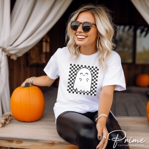 May include: White t-shirt with a black and white checkered square design featuring a cartoon ghost with a smiling face. The ghost is in the center of the square.