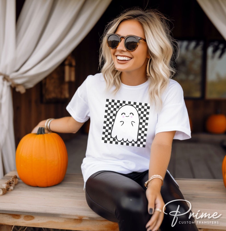 May include: White t-shirt with a black and white checkered square design featuring a cartoon ghost with a smiling face. The ghost is in the center of the square.