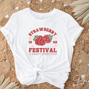 May include: White t-shirt featuring a retro design with the words "Strawberry Festival" in red and the numbers "1968". Two red strawberries with green leaves are in the center, with the phrase "A Berry Good Time" below.