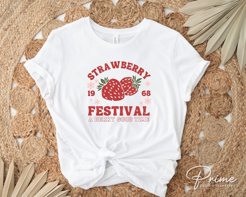 May include: White t-shirt featuring a retro design with the words "Strawberry Festival" in red and the numbers "1968". Two red strawberries with green leaves are in the center, with the phrase "A Berry Good Time" below.