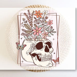 May include: A white skull with pink, peach, and green flowers growing out of the top. The skull is surrounded by a pink and white background with a brown border. Two butterflies are on either side of the skull.