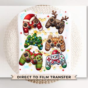 May include: A white t-shirt with a Christmas-themed design featuring six different video game controllers decorated with festive elements like holly, ornaments, and Santa hats. The text "Direct to Film Transfer" is printed at the bottom of the shirt.