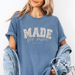 Puede incluir: Camiseta azul claro con la frase "MADE for more" en un dise&ntilde;o floral. El texto "Ephesians 2:10" est&aacute; impreso debajo. Las mangas est&aacute;n dobladas. La camiseta es de color azul claro.