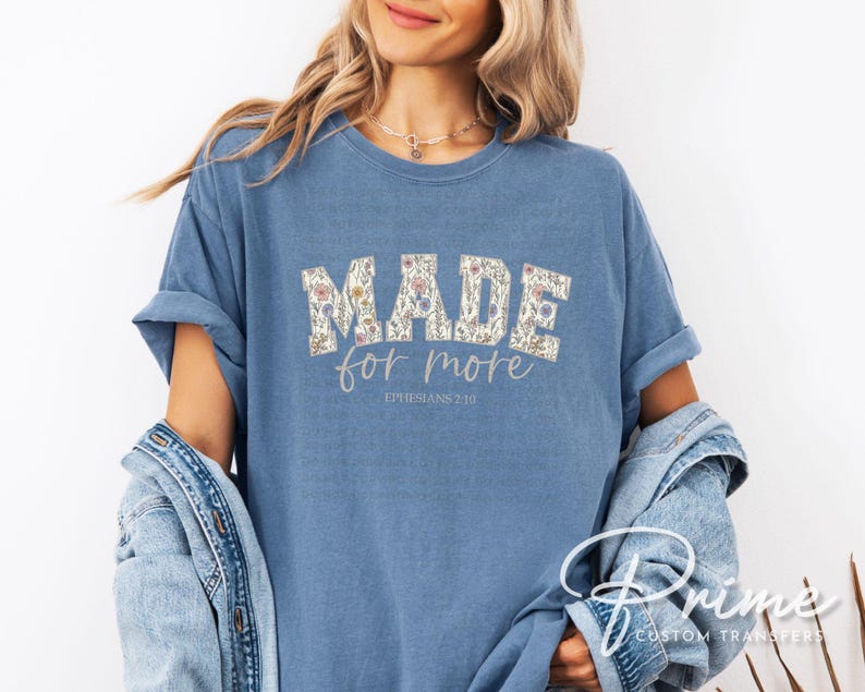 Puede incluir: Camiseta azul claro con la frase "MADE for more" en un dise&ntilde;o floral. El texto "Ephesians 2:10" est&aacute; impreso debajo. Las mangas est&aacute;n dobladas. La camiseta es de color azul claro.