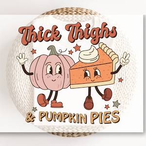 K&ouml;nnte beinhalten: Ein wei&szlig;es T-Shirt mit dem Text "Thick Thighs & Pumpkin Pies" in einer Retro-Schrift. Der Text ist von einem rosa K&uuml;rbis und einem St&uuml;ck K&uuml;rbiskuchen mit Gesichtern und Armen umgeben.