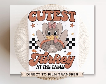 May include: A white t-shirt with a cute cartoon turkey wearing a bow. The text "Cutest Turkey at the Table" is printed in brown and orange letters. The shirt is for direct to film transfer.