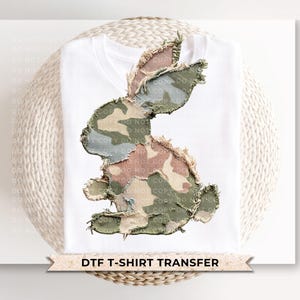 May include: White t-shirt featuring a camouflage bunny design. The bunny is made of various fabric patches in shades of green, brown, and pink. The text "DTF T-SHIRT TRANSFER" is at the bottom.