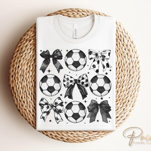 Puede incluir: Camiseta blanca con un dise&ntilde;o de bal&oacute;n de f&uacute;tbol y lazo en blanco y negro. El dise&ntilde;o incluye seis balones de f&uacute;tbol y seis lazos con varios patrones. La camiseta se muestra sobre una superficie circular tejida.