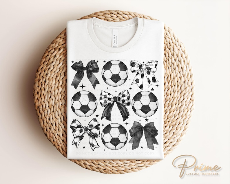 Puede incluir: Camiseta blanca con un dise&ntilde;o de bal&oacute;n de f&uacute;tbol y lazo en blanco y negro. El dise&ntilde;o incluye seis balones de f&uacute;tbol y seis lazos con varios patrones. La camiseta se muestra sobre una superficie circular tejida.