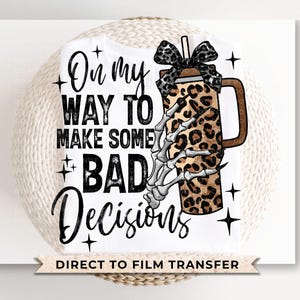 May include: White t-shirt with the text "On my way to make some bad decisions." A leopard print coffee cup with a skeleton hand and a black bow is on the right side of the shirt. The words "Direct to Film Transfer" are at the bottom.