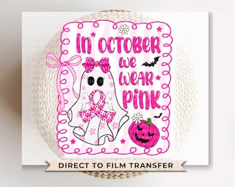 May include: A pink and white graphic with the text "In October We Wear Pink." The design features a ghost wearing a pink ribbon, a pink pumpkin, and bats. The words "Direct to Film Transfer" are at the bottom.