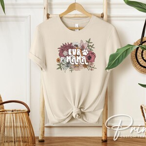 May include: A cream-colored t-shirt with the text "FUR MAMA" in a floral design. The design includes a paw print and various flowers in shades of pink, burgundy, and white. The shirt is hanging on a wooden hanger.