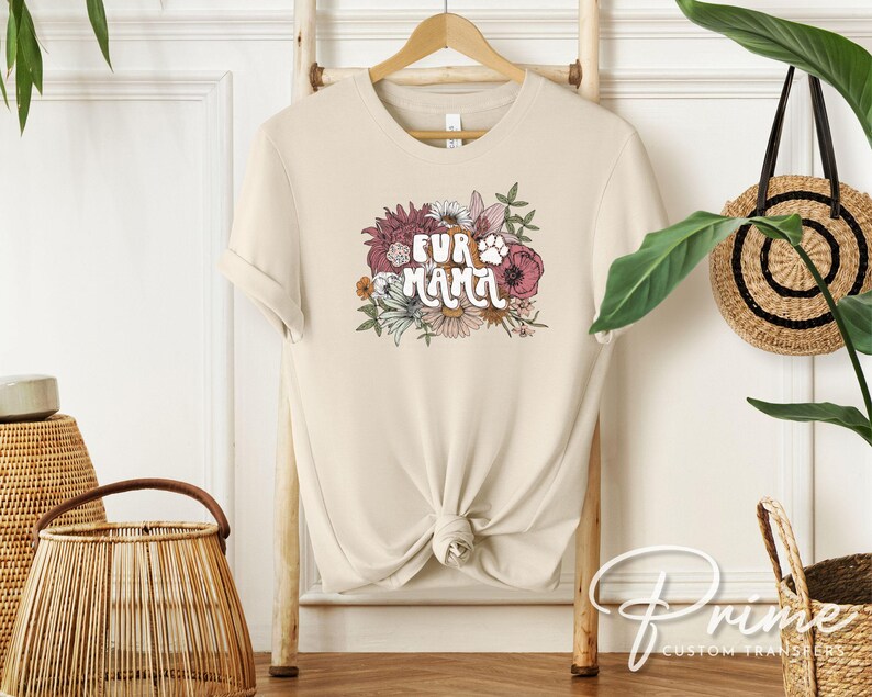 May include: A cream-colored t-shirt with the text "FUR MAMA" in a floral design. The design includes a paw print and various flowers in shades of pink, burgundy, and white. The shirt is hanging on a wooden hanger.
