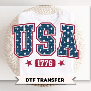 Op de afbeelding: Wit T-shirt met het woord "USA" in verweerde rode en blauwe letters met een sterrenpatroon. Onder het woord "USA" staat het nummer "1776" in een rode rechthoek. De tekst "DTF TRANSFER" staat onderaan.