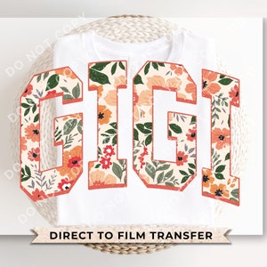 May include: White t-shirt with the word "GIGI" in large, bold letters. The letters are filled with a floral pattern featuring orange, pink, and green flowers. The text "DIRECT TO FILM TRANSFER" is printed below the letters.