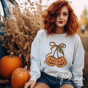 May include: A light blue sweatshirt featuring a Halloween-themed design of two jack-o'-lanterns tied together with an orange bow. Two orange pumpkins are in the foreground. The sweatshirt is part of a fall-themed collection.